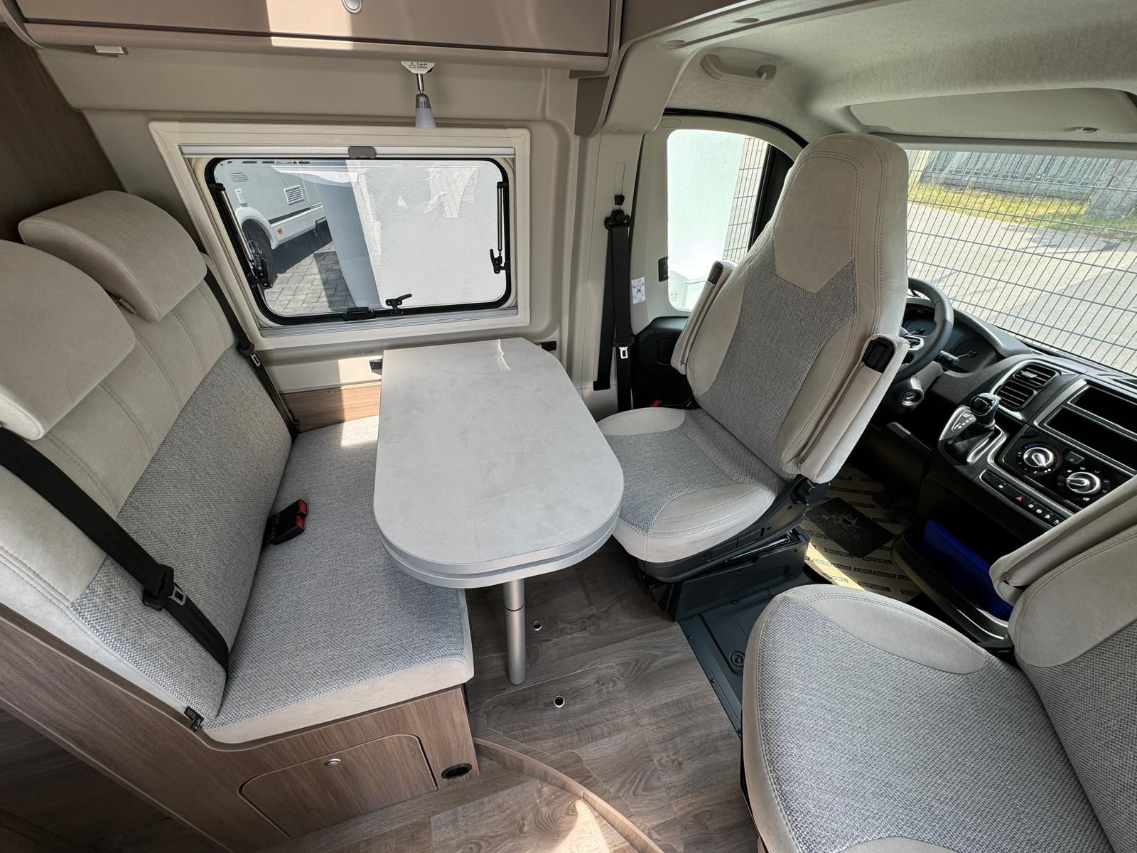 Innenraum eines Wohnmobils mit Sitzen, Tisch und Fenster. Hellgraue und neutrale Töne dominieren den Raum.