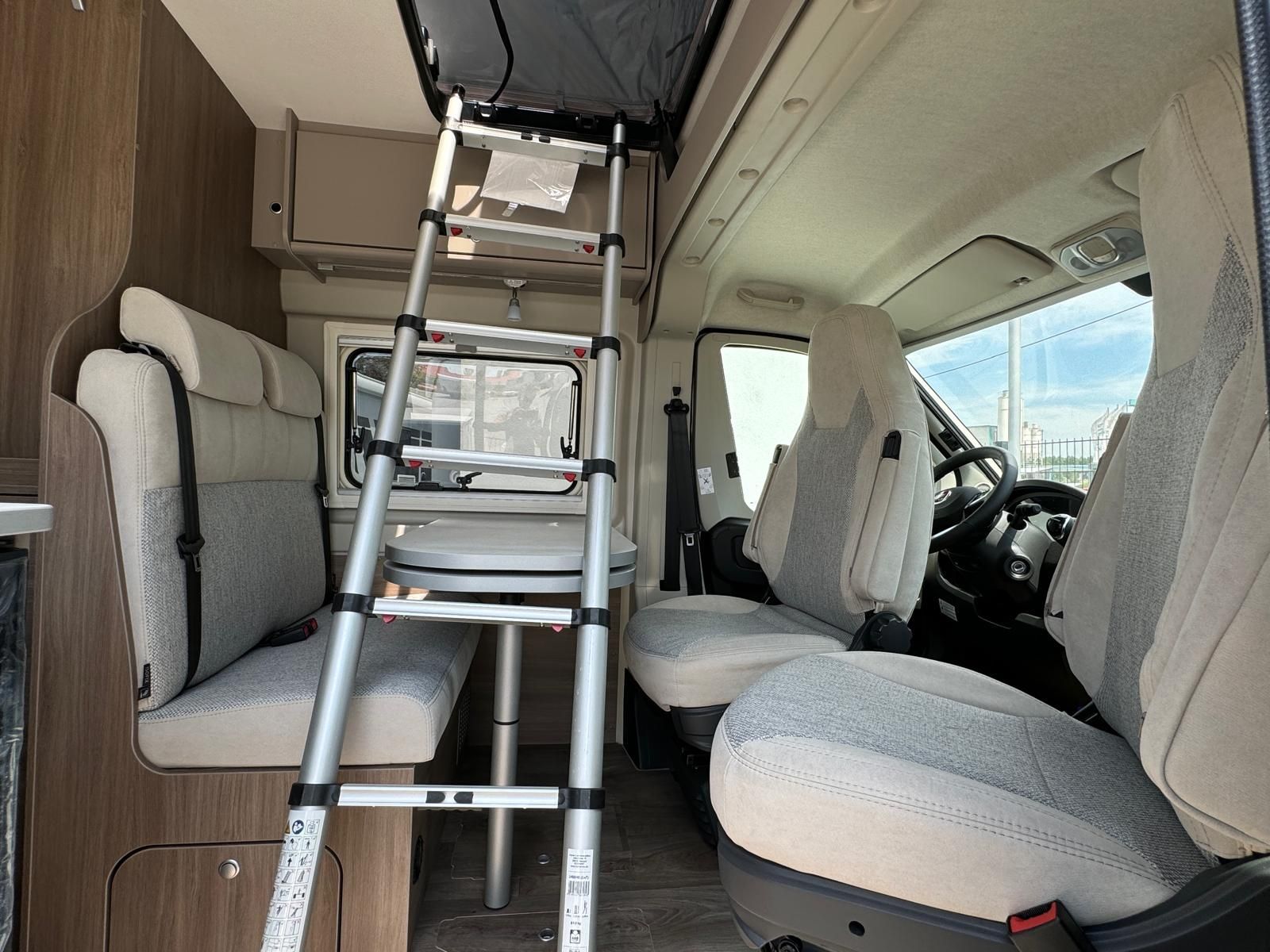 Innenraum eines Wohnmobils mit Sitzgelegenheiten, einer Leiter und Stauraum. Grau- und Beigetöne dominieren.