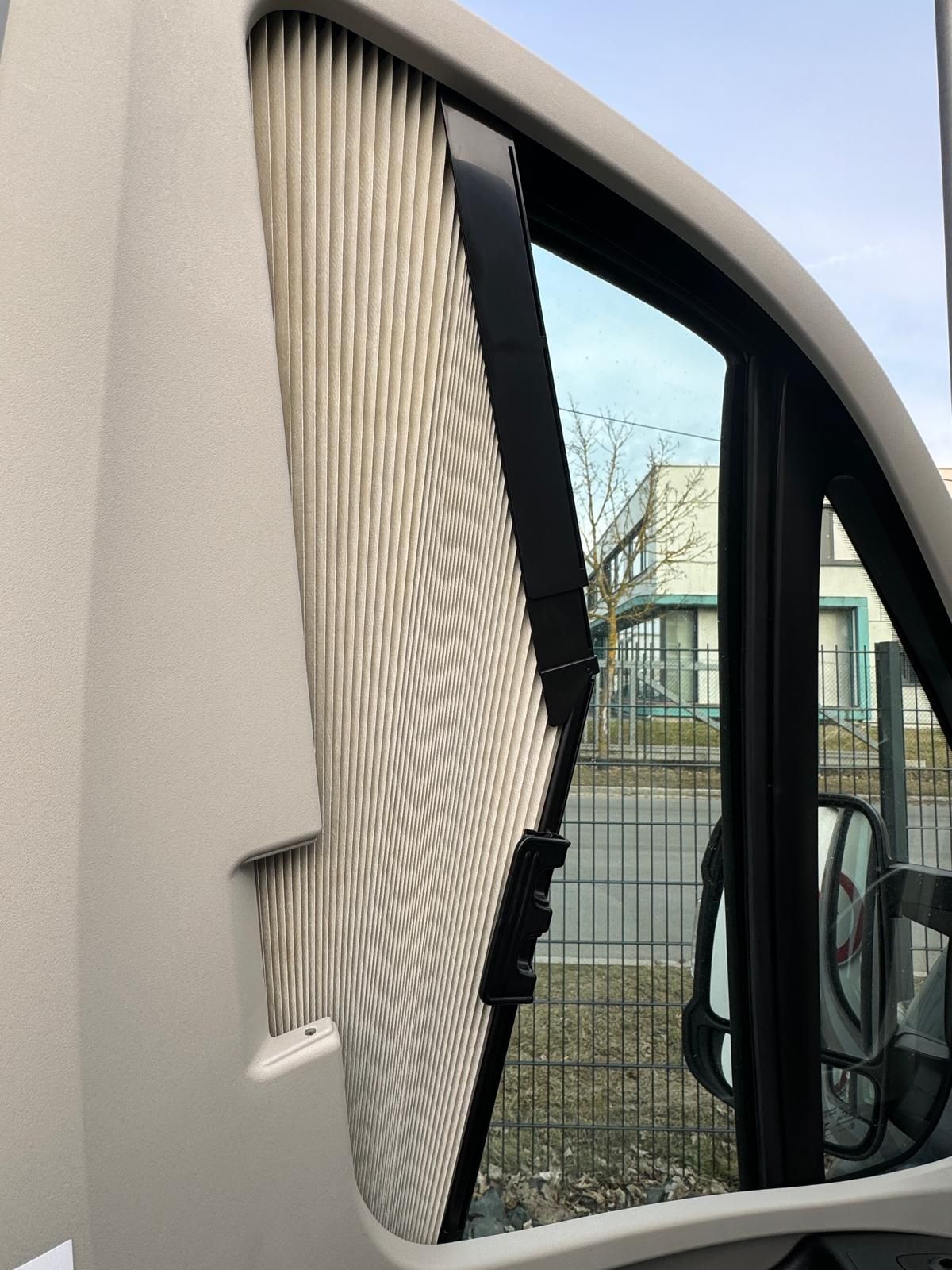 Wohnmobilfenster mit teilweise heruntergelassenen Plissees, die den Blick auf eine Straße, einen Zaun und Gebäude freigeben.