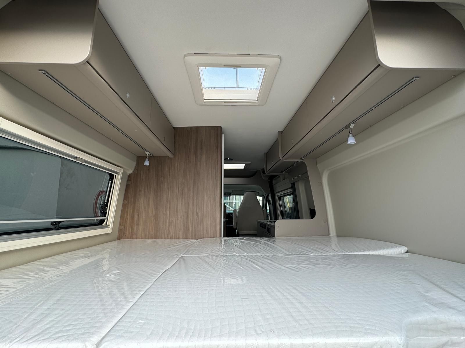 Innenansicht eines Wohnmobils mit Bett, Staufach über dem Kopf, Dachfenster und offener Tür.