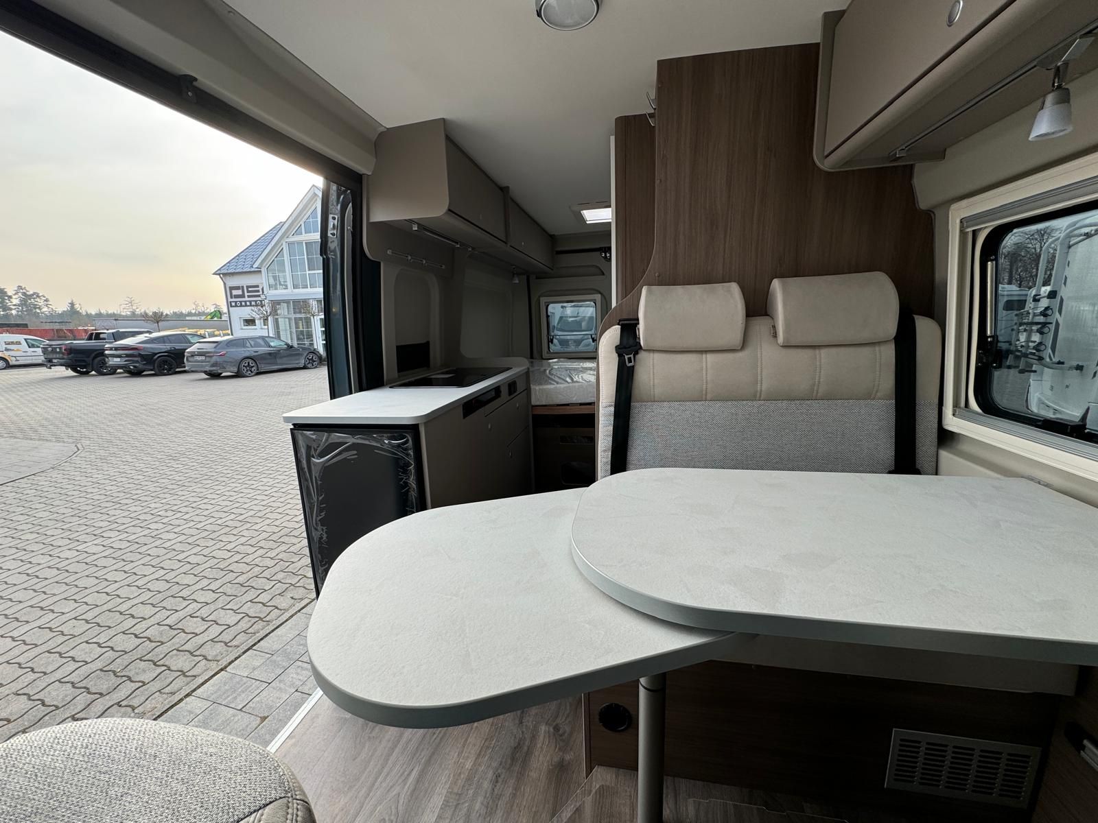 Innenansicht eines Wohnmobils mit Tisch, Sitzgelegenheiten, Küchenbereich und Blick nach draußen.