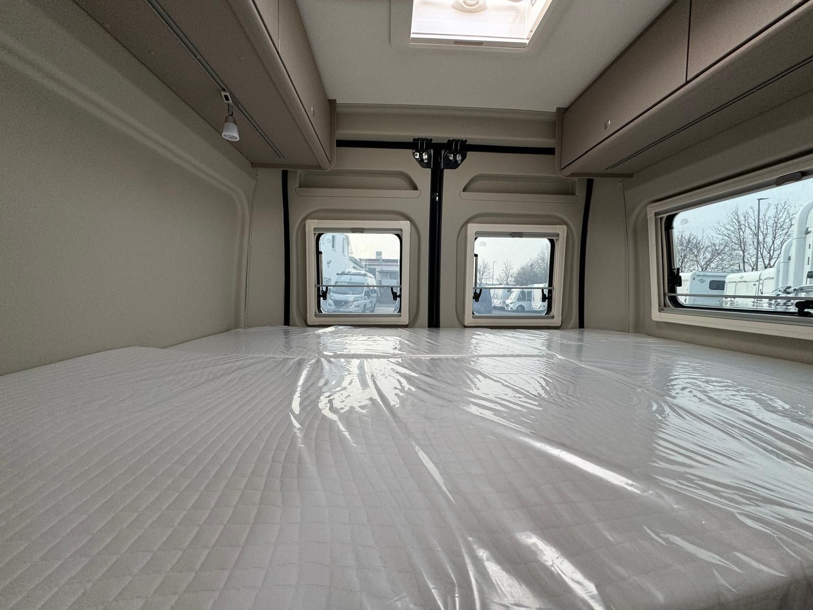 Innenraum eines Wohnmobils mit einem großen, mit Plastikfolie abgedeckten Bett, drei Fenstern und Staufächern über dem Kopf.