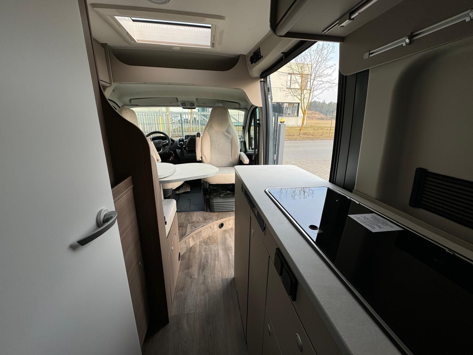 Innenansicht eines Wohnmobils mit Küche und Bett. Heller Holzboden und Wände in neutralen Farbtönen.