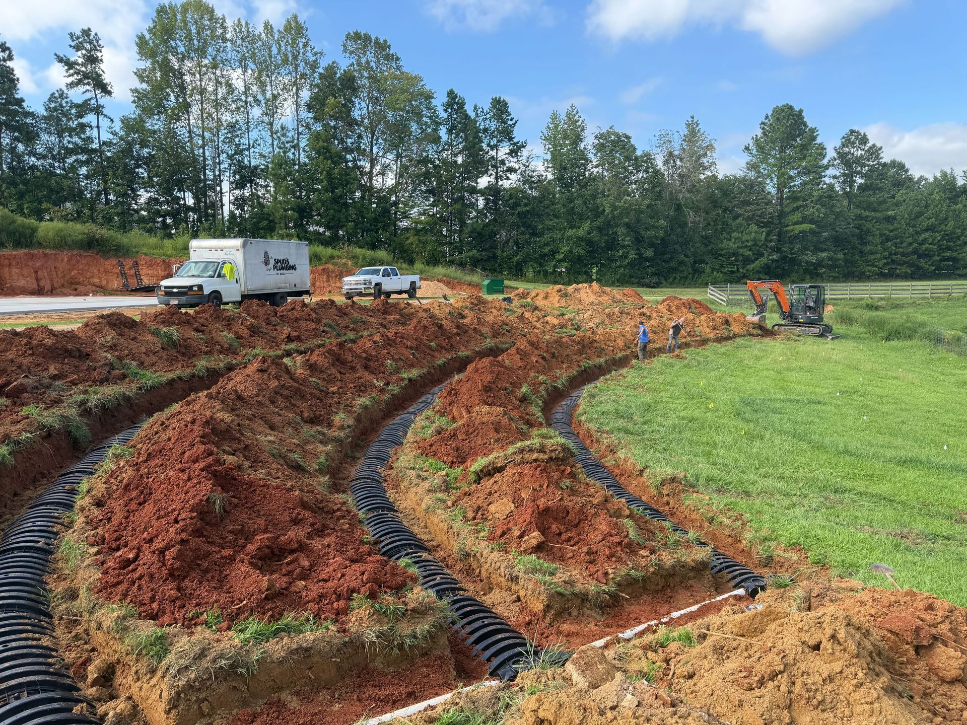 A construction site showing two curved trenches containing black plastic septic drainage chambers in red soil.