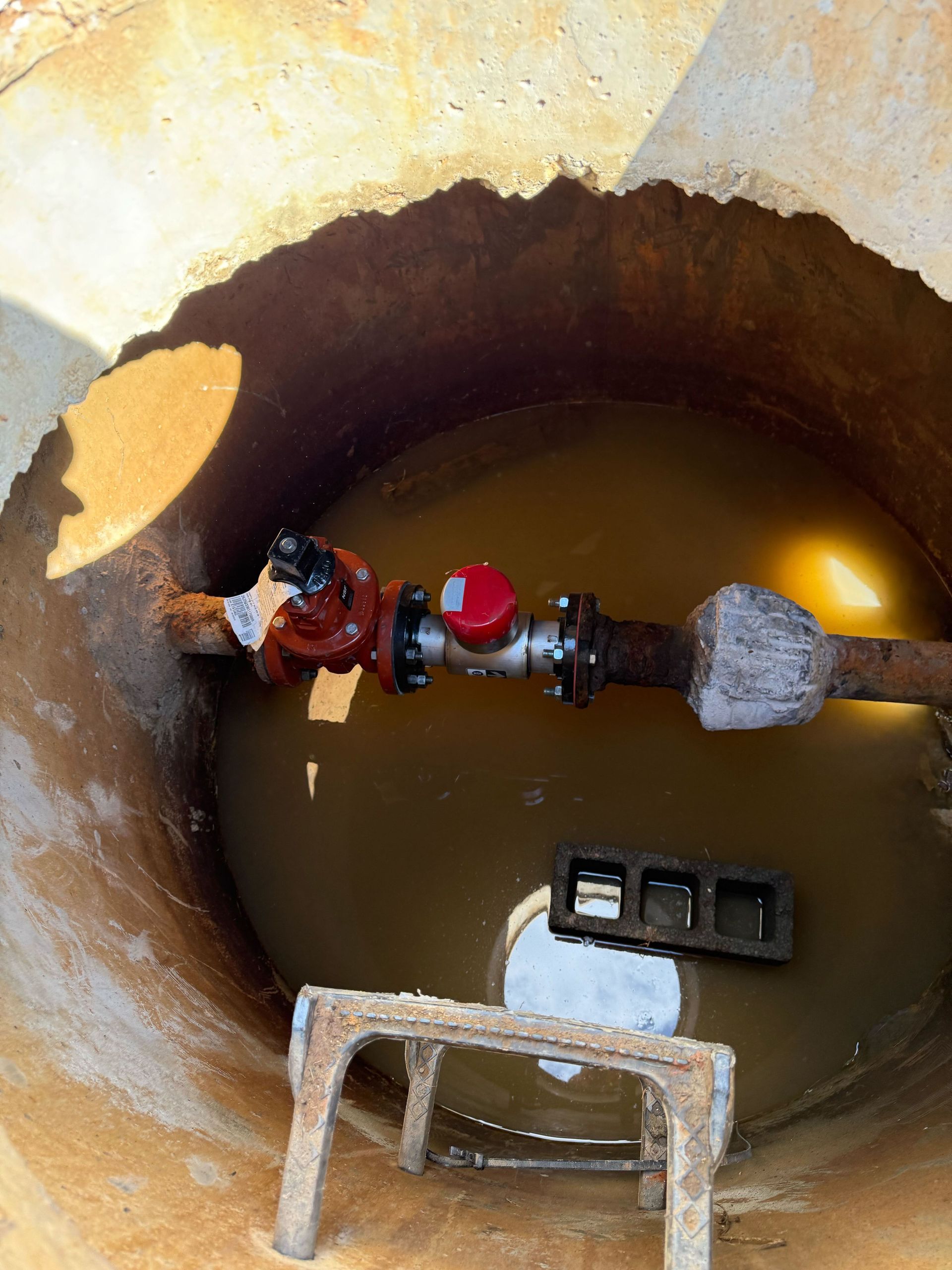 A metal pipe with a red valve and meter sitting in a pool of murky water inside a circular concrete manhole.