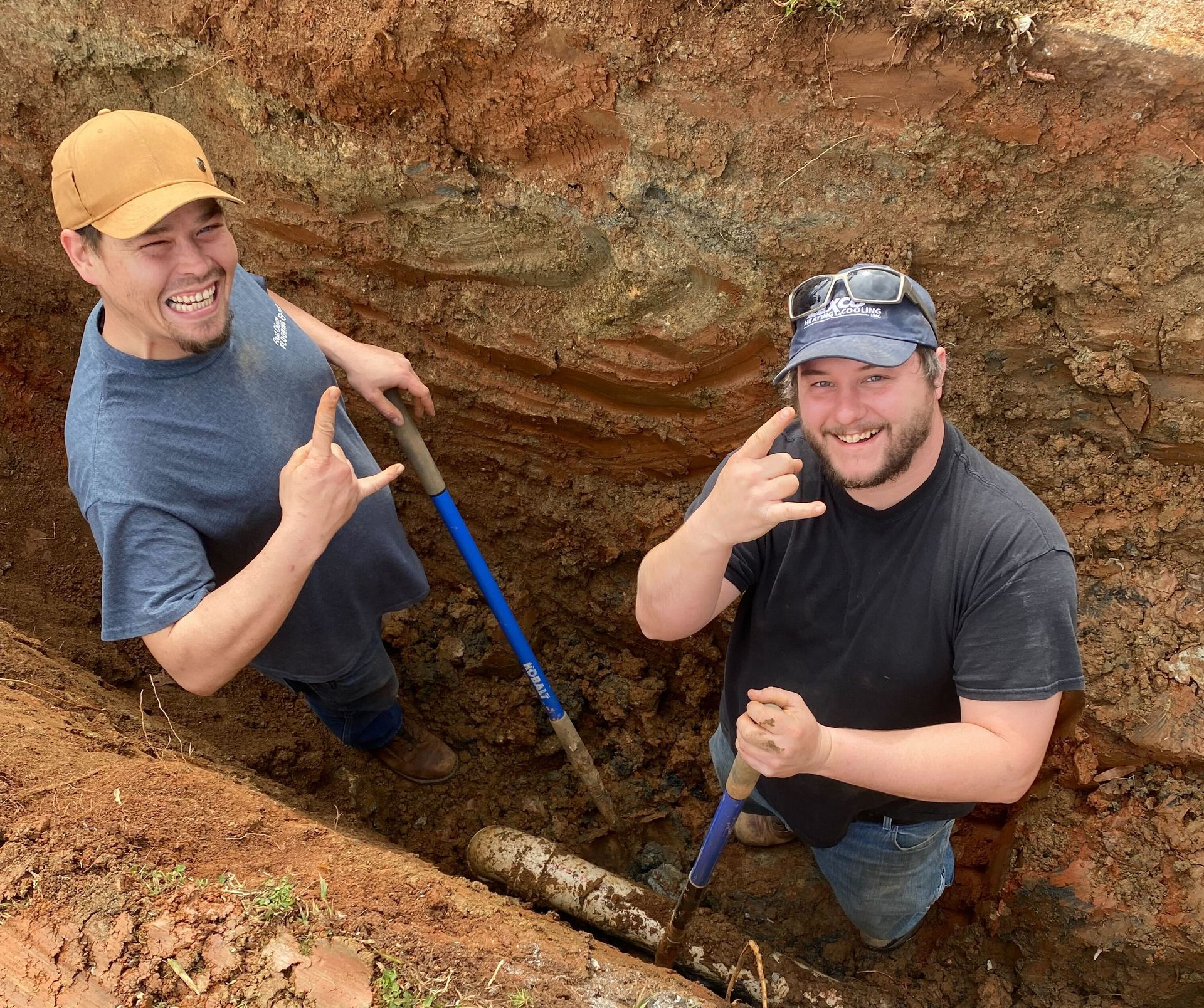 Two people smile and make hand gestures while standing in a deep dirt trench, each holding a shovel near a pipe.