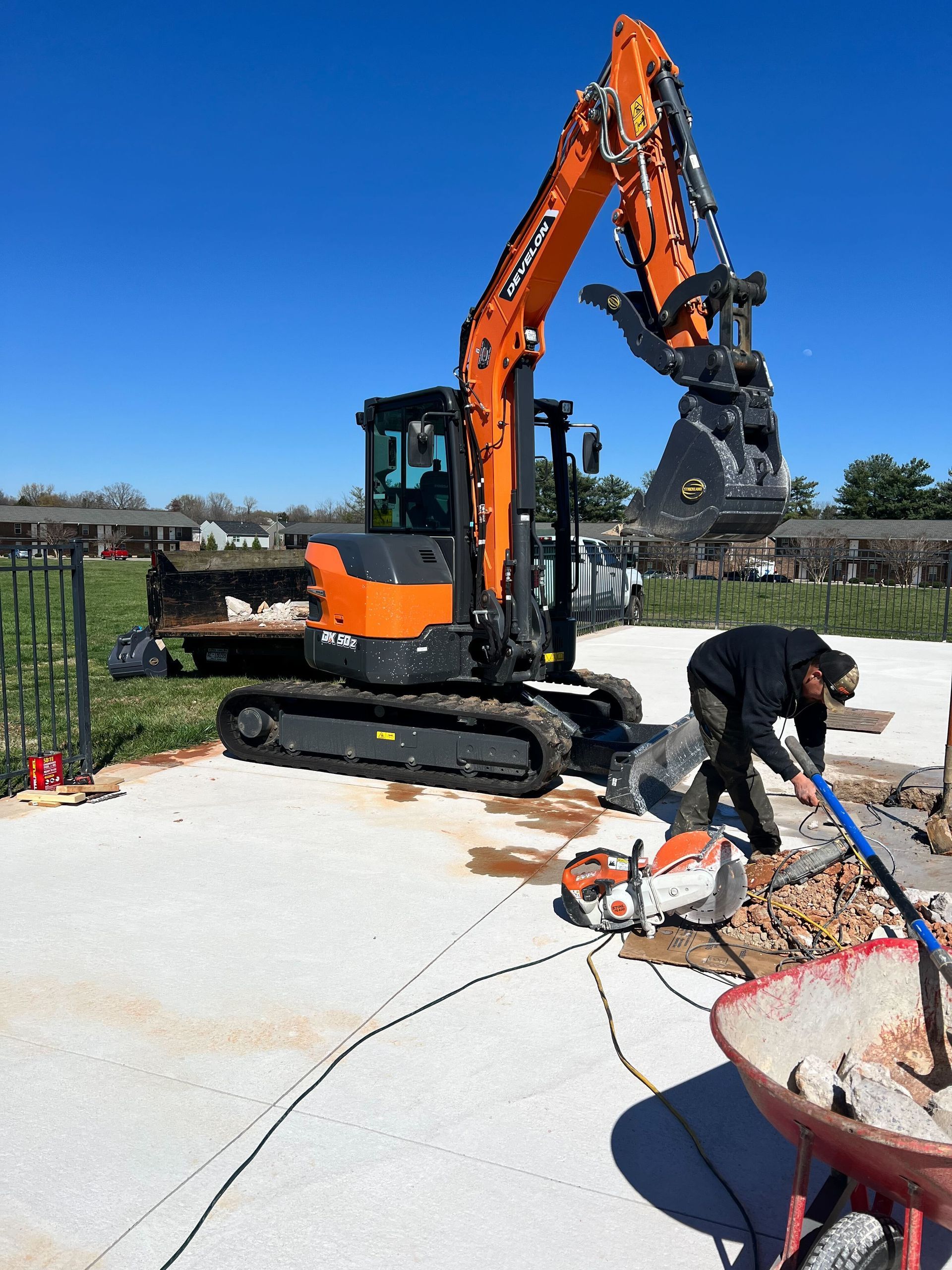 A worker uses a concrete saw near a construction site with an orange excavator in the background.