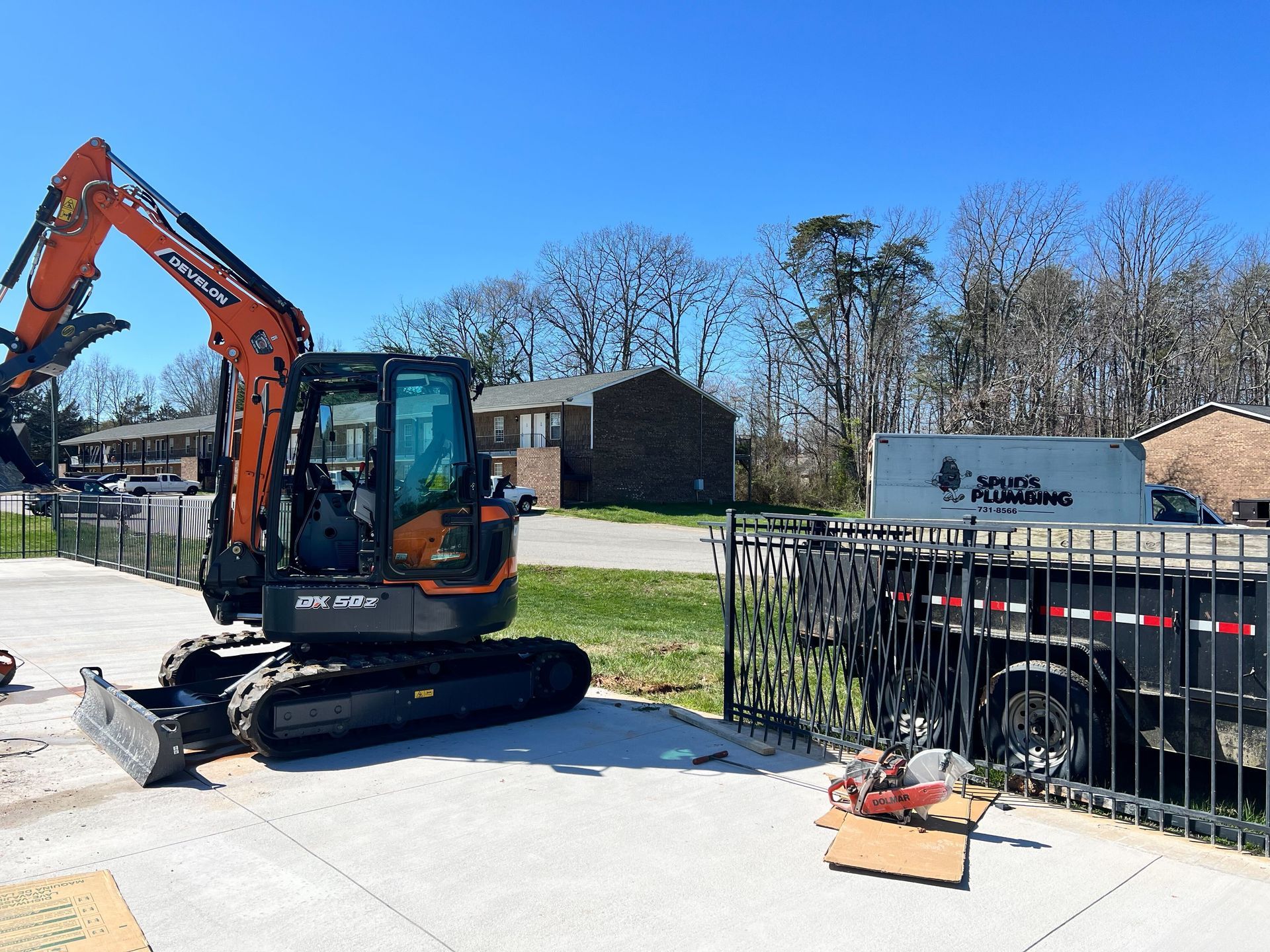 An orange excavator sits on a concrete pad next to a black utility trailer marked 
