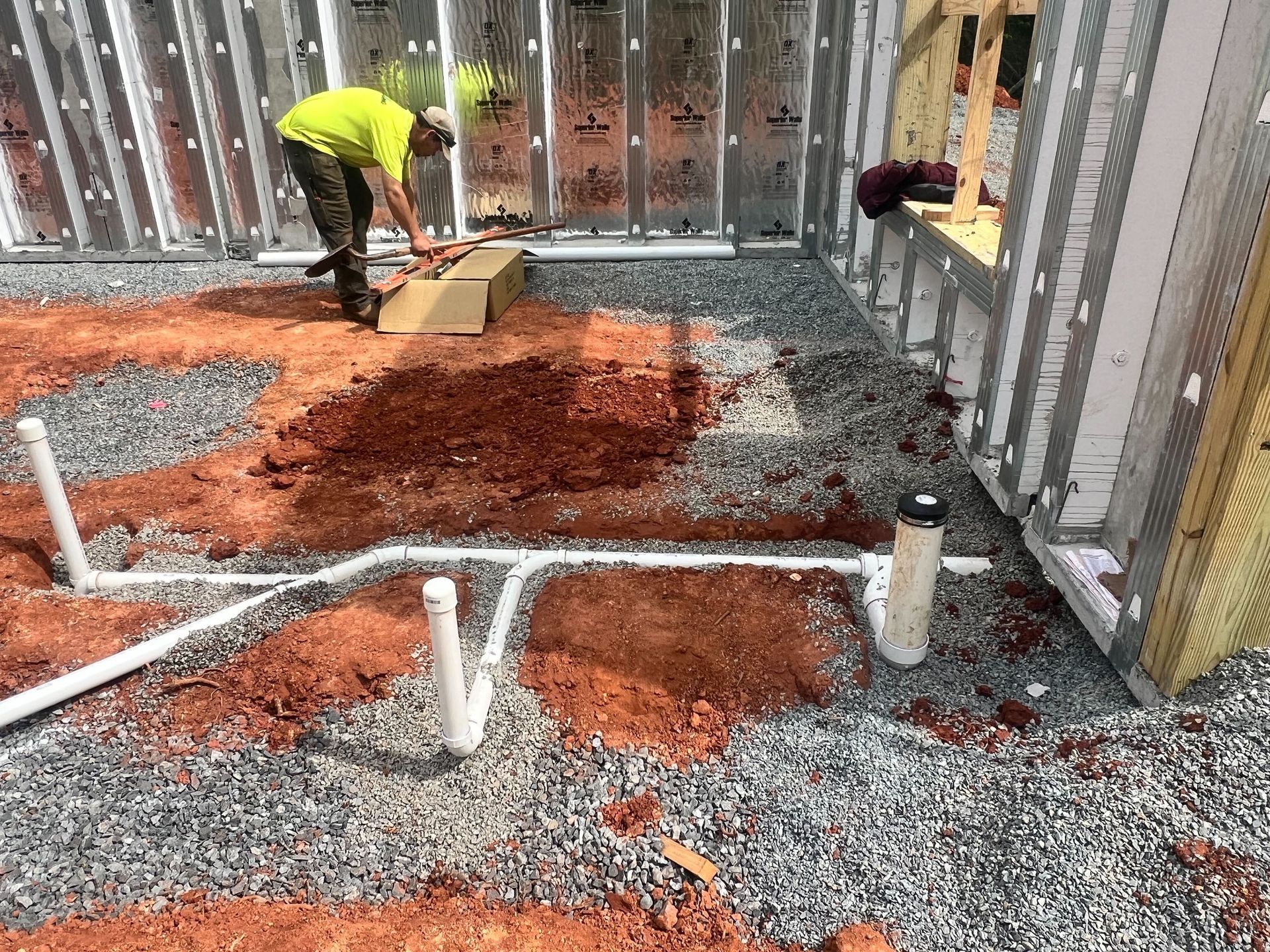 A worker in a yellow shirt stands in a construction site with white PVC pipes laid out on a gravel and dirt ground.