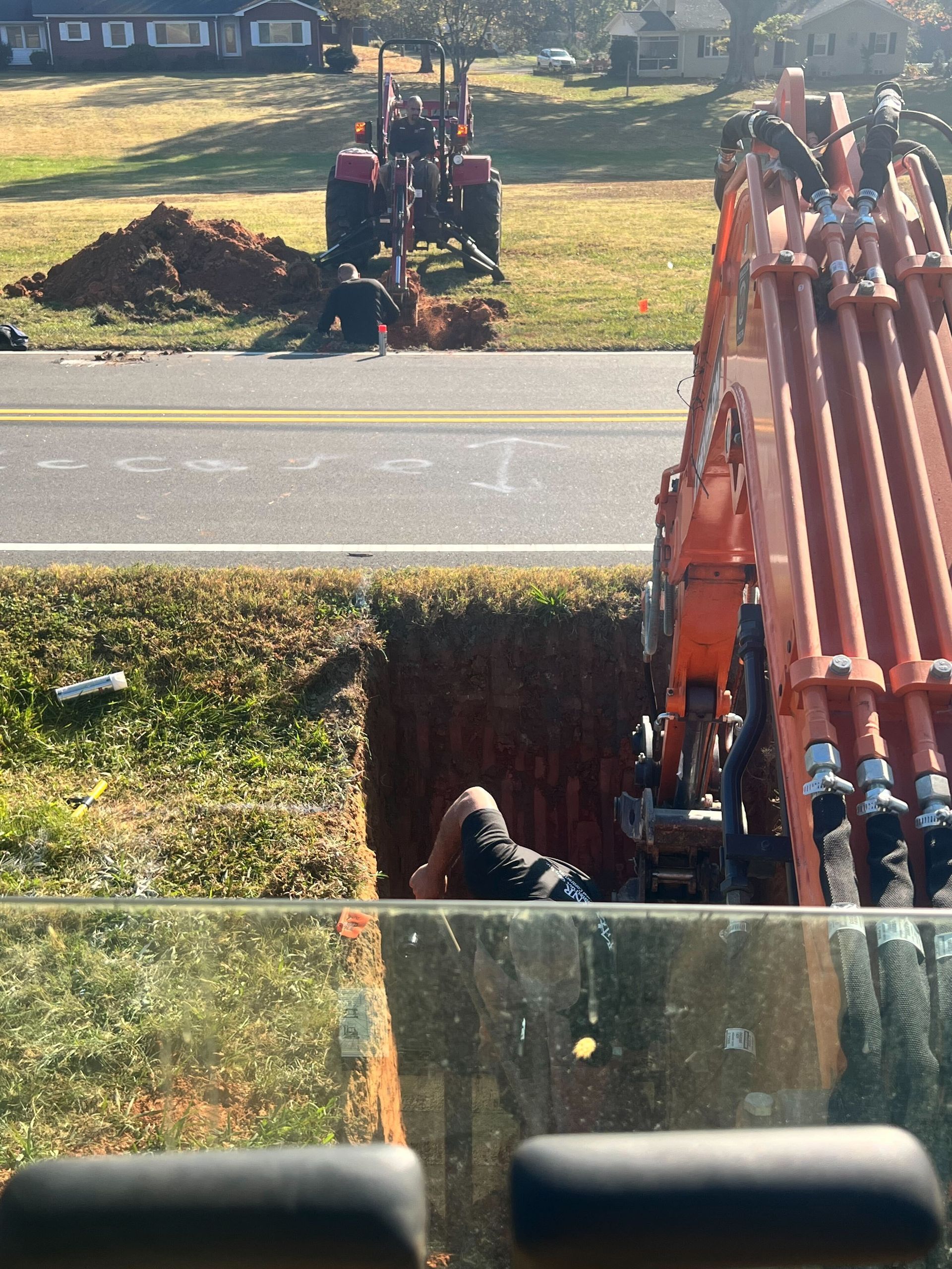 An excavator and a tractor work on a roadside utility project with workers in a trench and near a dirt pile.