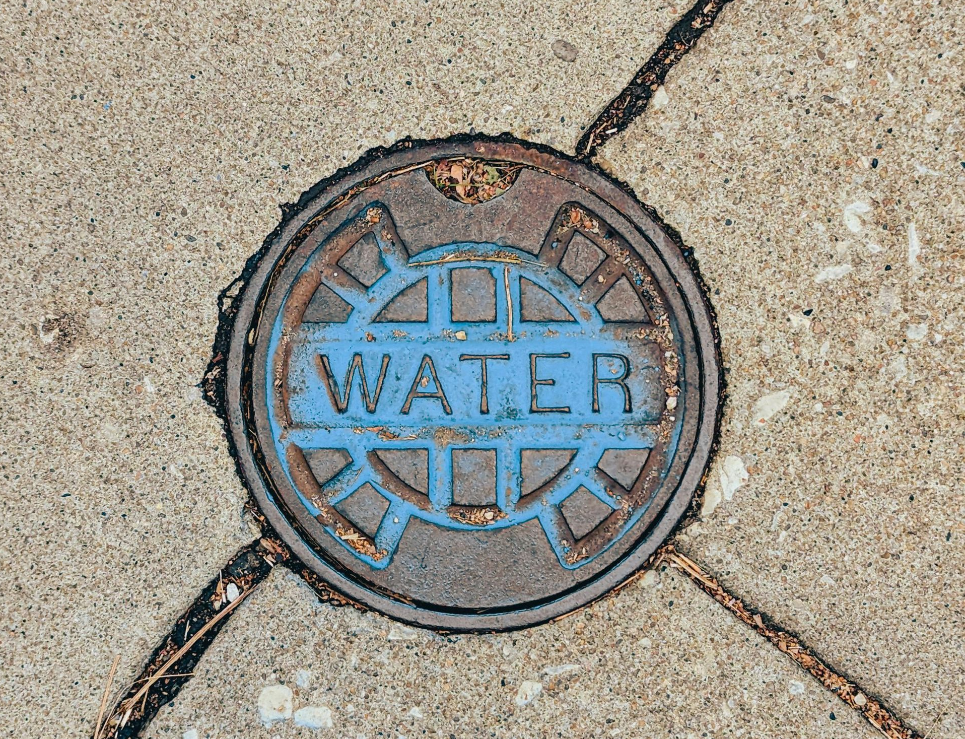 Blue water utility cover set in a tan concrete sidewalk with diagonal cracks.