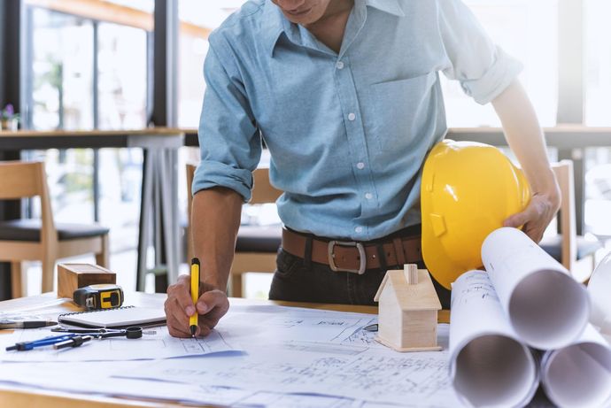 A man sits at a table holding a hard hat and writing on a blueprint.