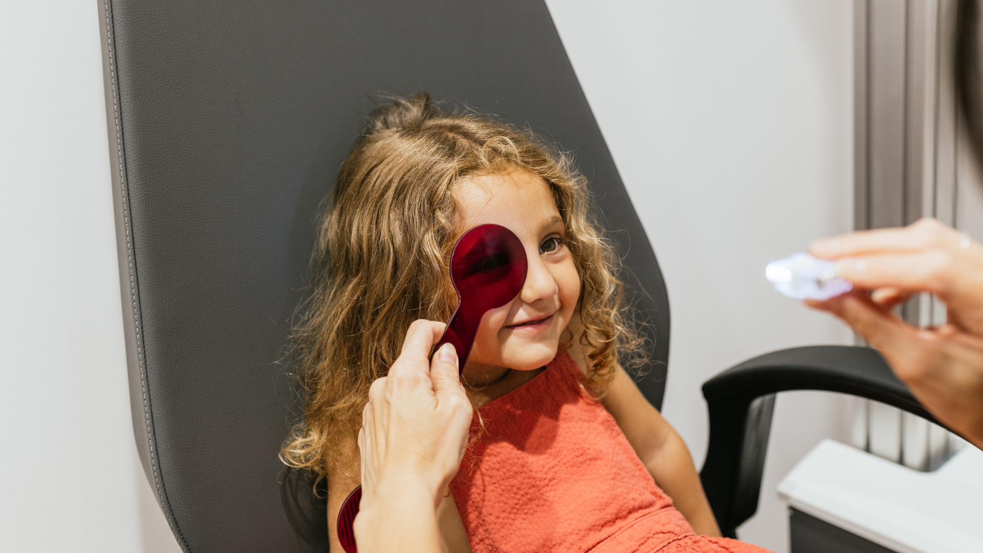 Eye exam for a smiling child using a handheld occluder in a clinic chair