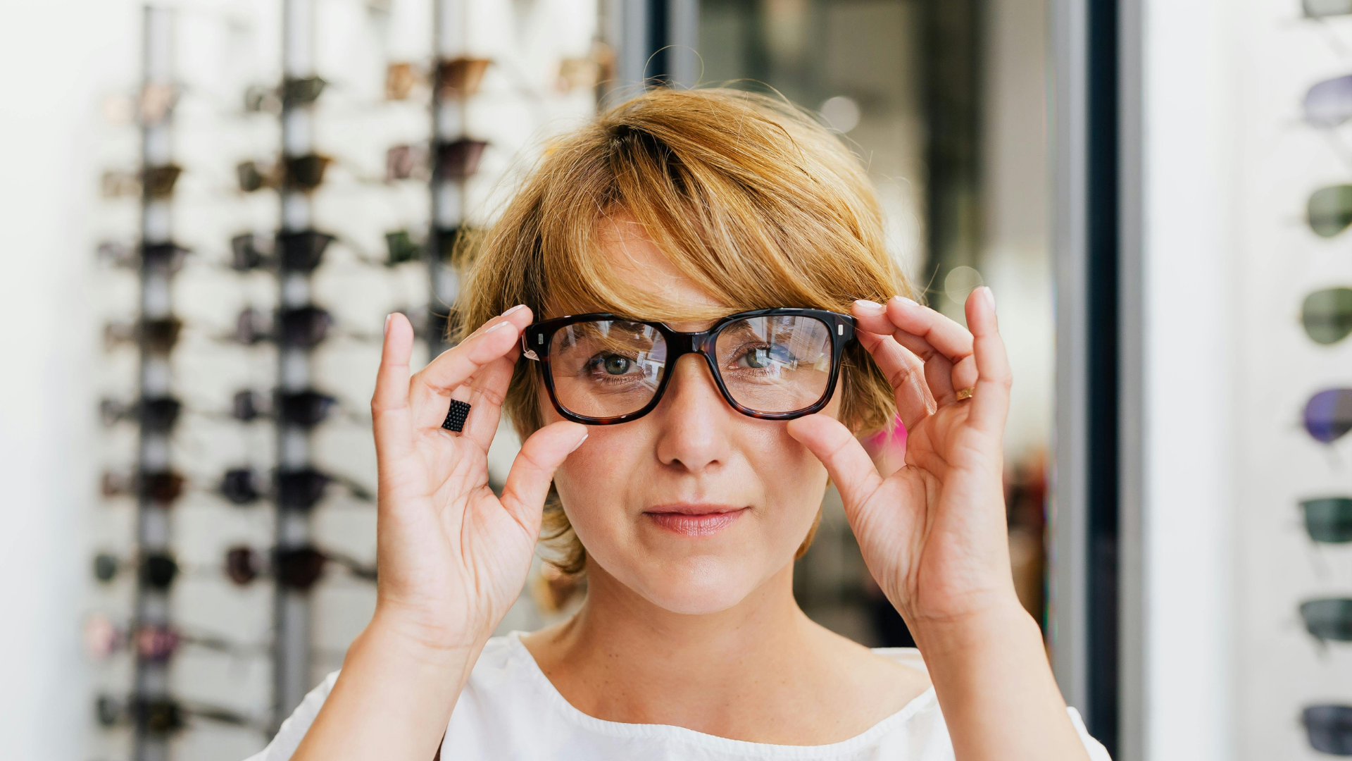 Woman adjusting large black eyeglasses in an eyewear store, with rows of glasses in the background