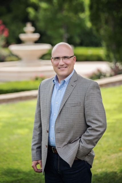 Bald man in blazer, blue shirt, and glasses, standing outdoors in front of a fountain, smiling.