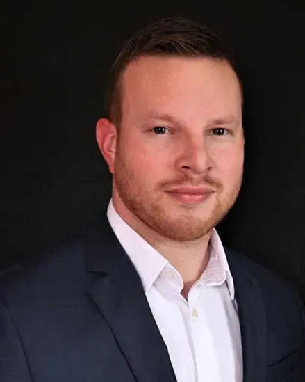 Man in suit smiling, with short brown hair, against a black background.