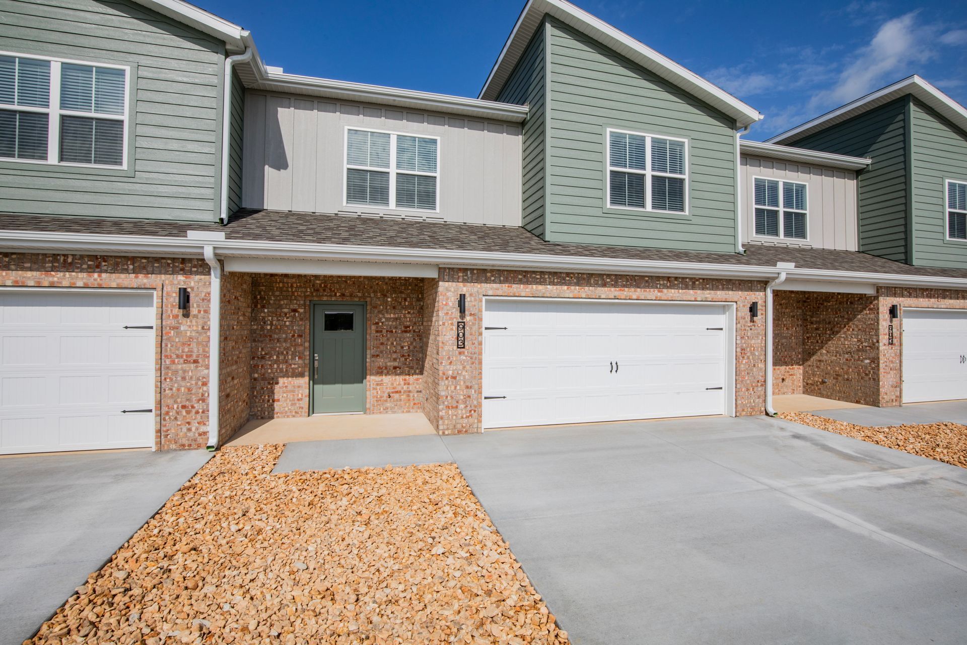 Row of townhouses with attached garages, green and tan siding, brick facade, and gravel walkway.