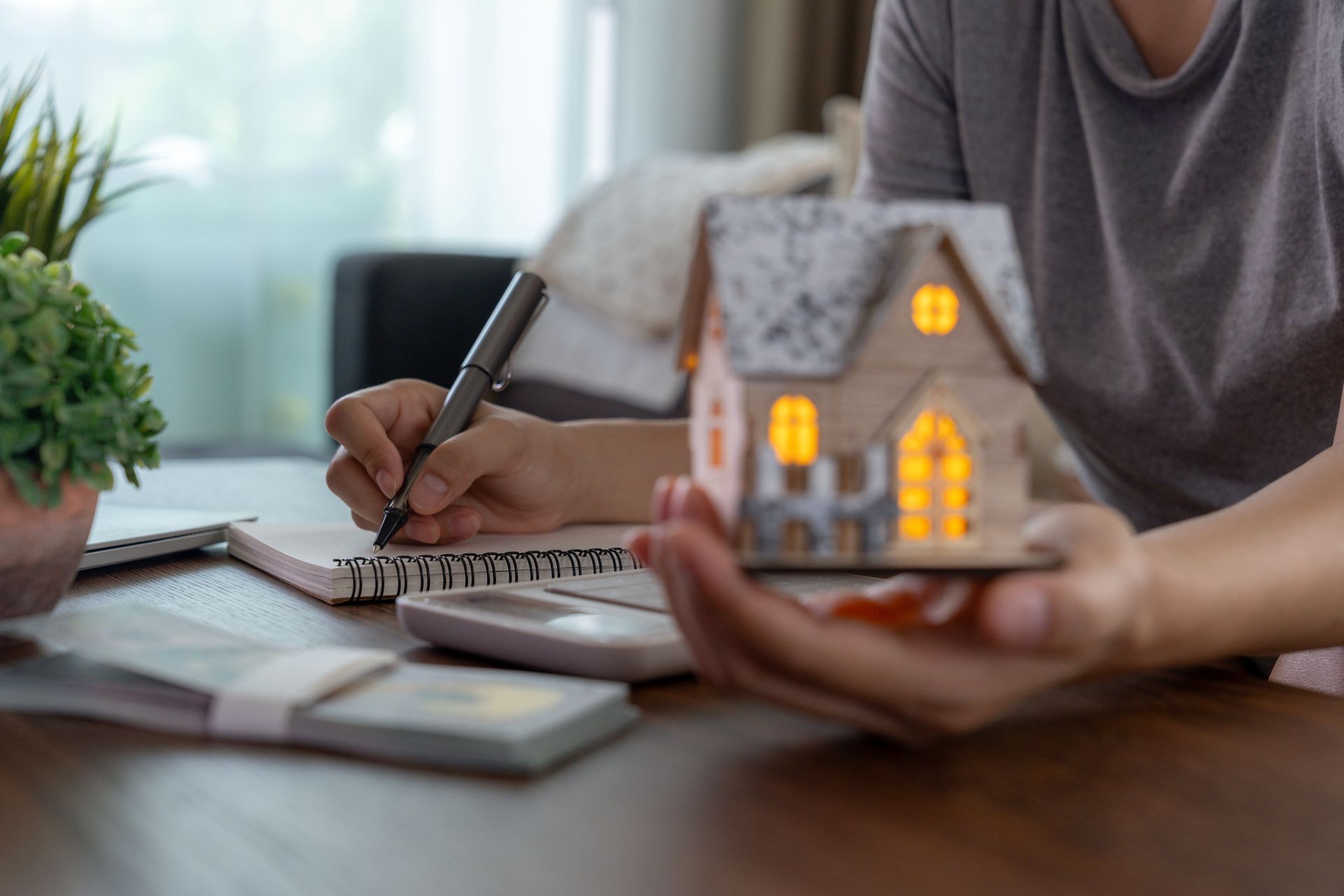 Person writing in notebook, holding miniature house, money and calculator on desk.