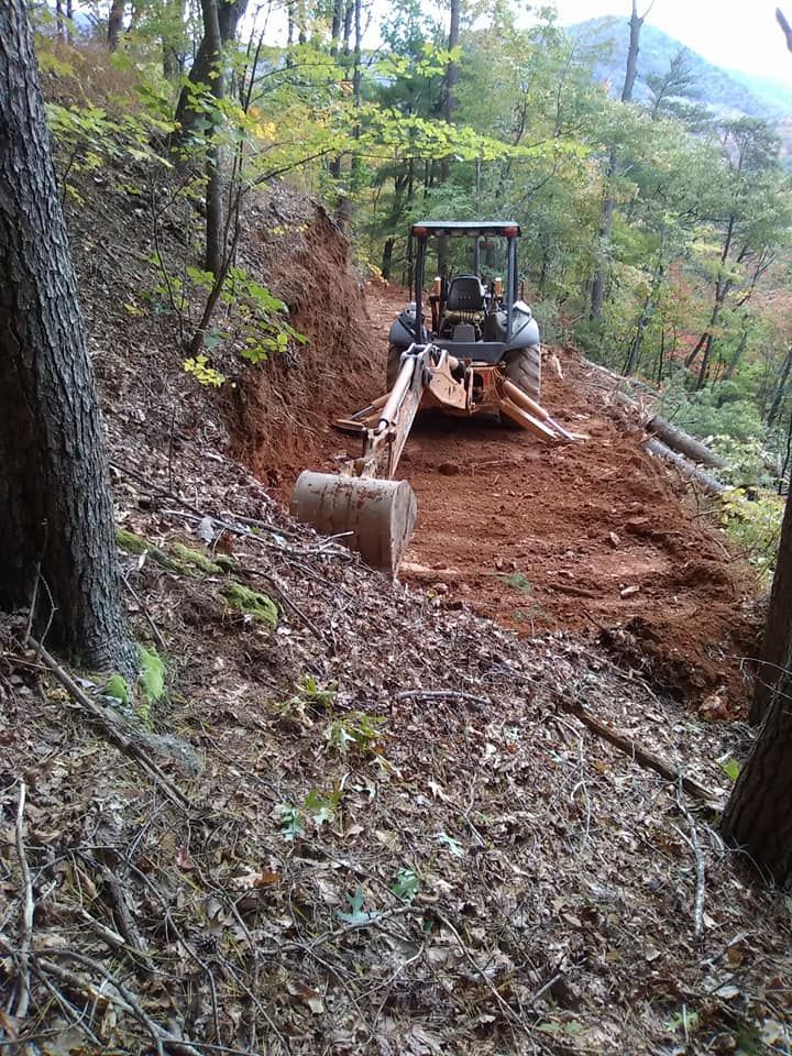 A backhoe clearing a dirt trail on a hillside in a wooded area.