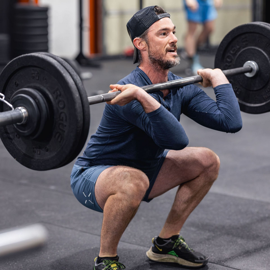 Man performing a front squat with a barbell in a gym, wearing a cap and navy workout clothes.