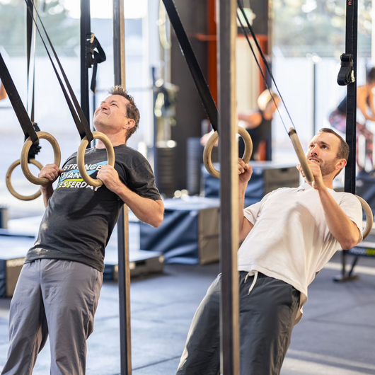Two people doing ring rows in a bright gym, leaning back while gripping gymnastic rings.