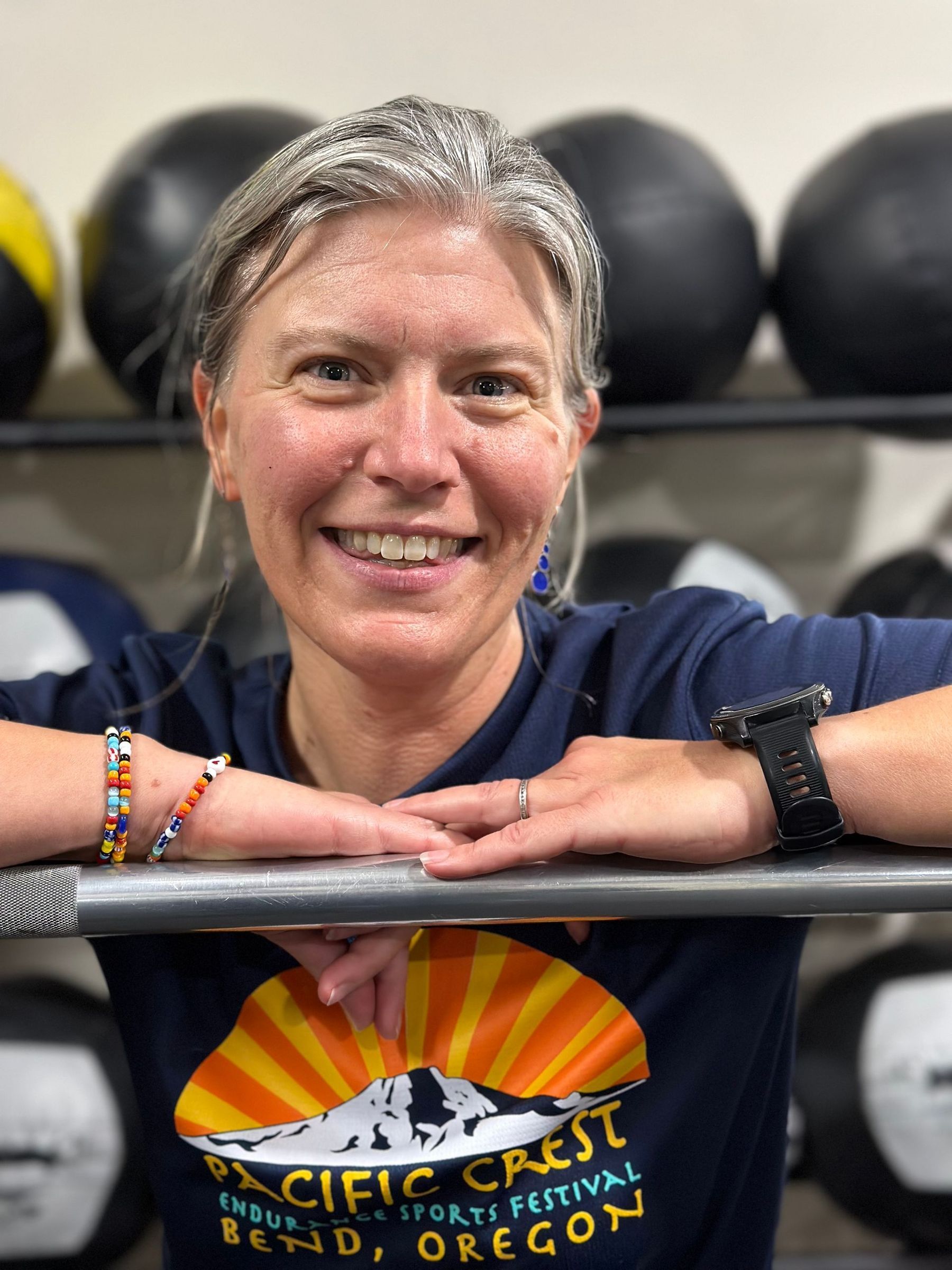 Smiling person leaning on a barbell rack in a gym, with medicine balls in the background.