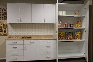 A kitchen with white cabinets , drawers , and a pantry filled with cereal.
