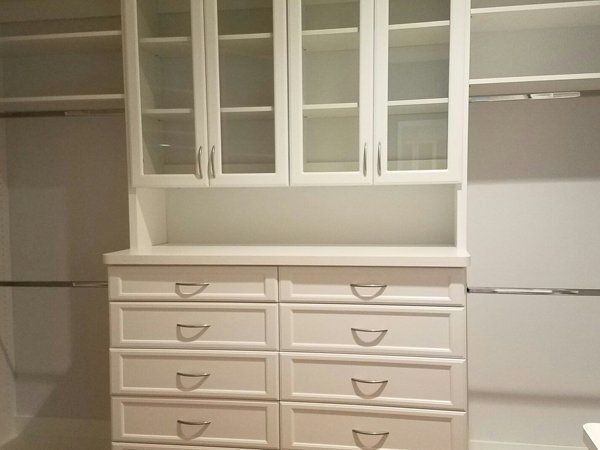 A walk in closet with white drawers and glass doors.