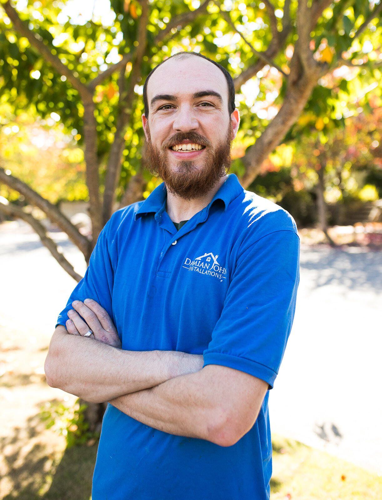 A man with a beard wearing a blue shirt is standing with his arms crossed in front of a tree.