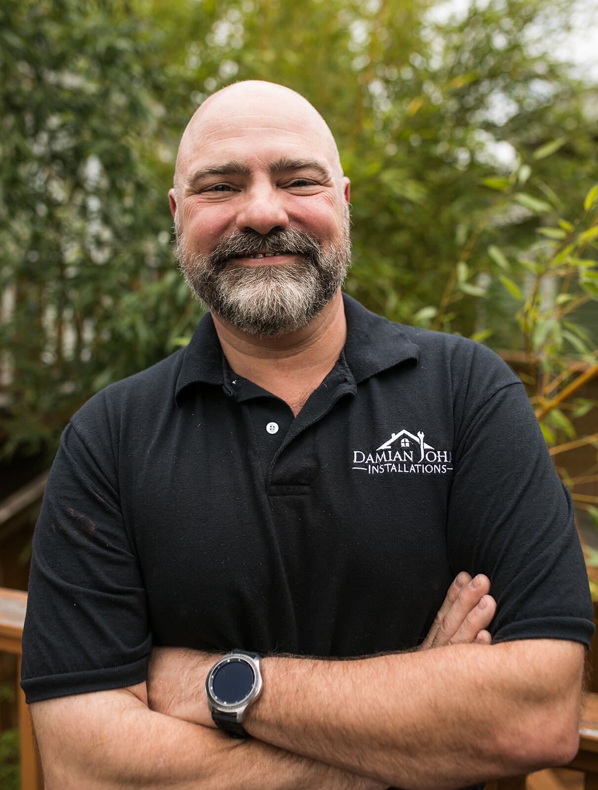 A bald man with a beard and a watch is standing with his arms crossed and smiling.