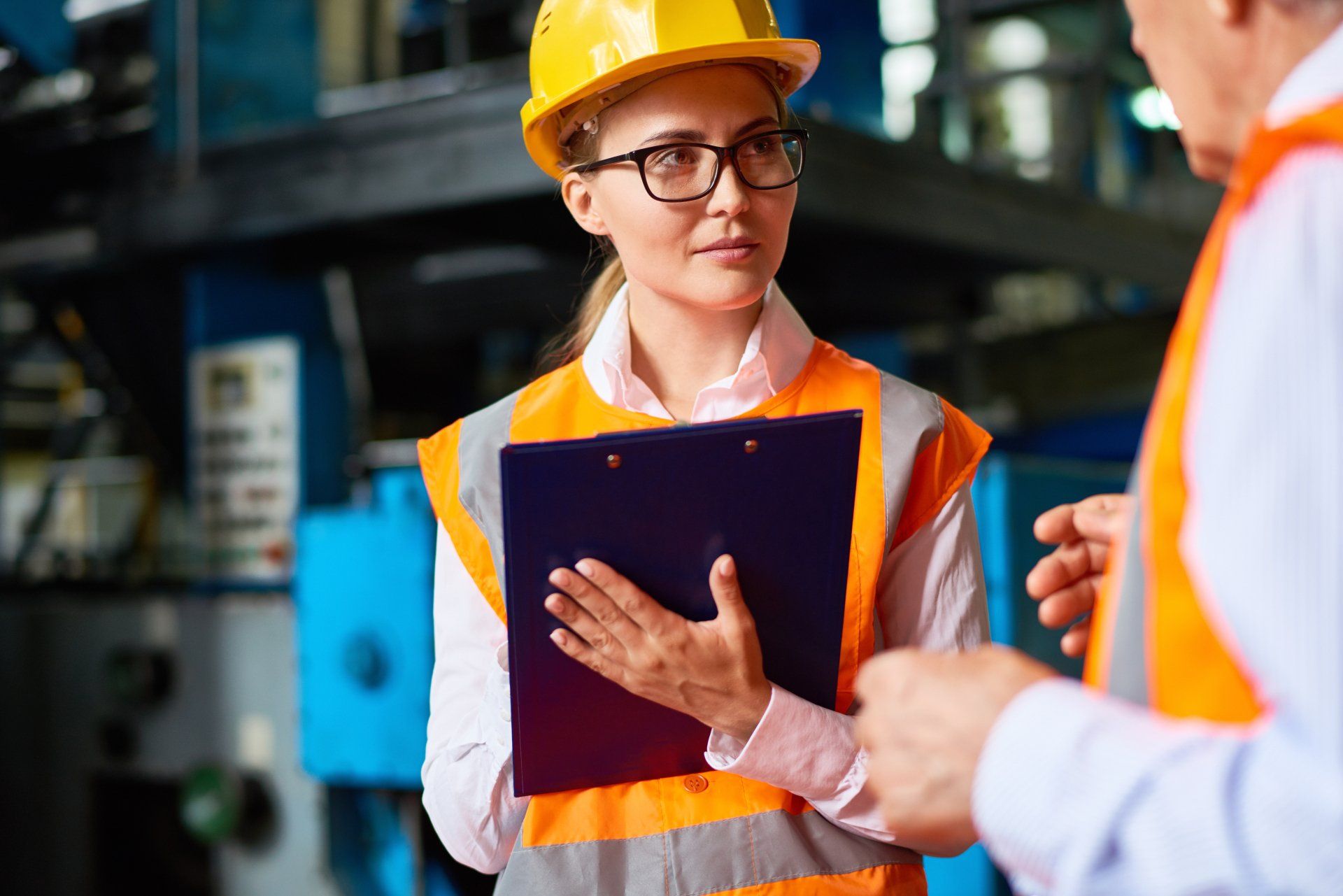 A woman in a hard hat is holding a clipboard and talking to a man in a factory.