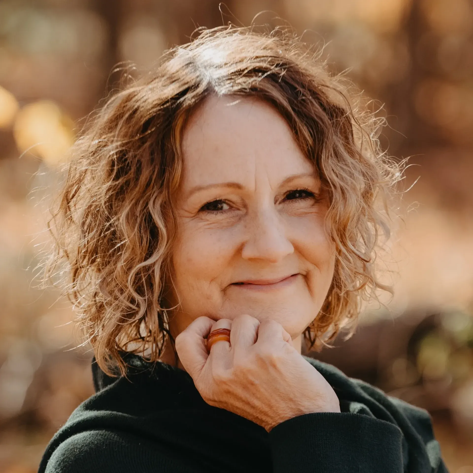 A woman with curly hair is smiling and holding her hand to her chin.