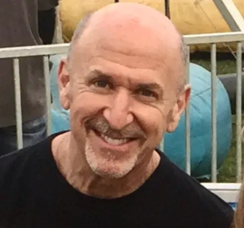 A bald man with a beard is smiling in front of a fence.