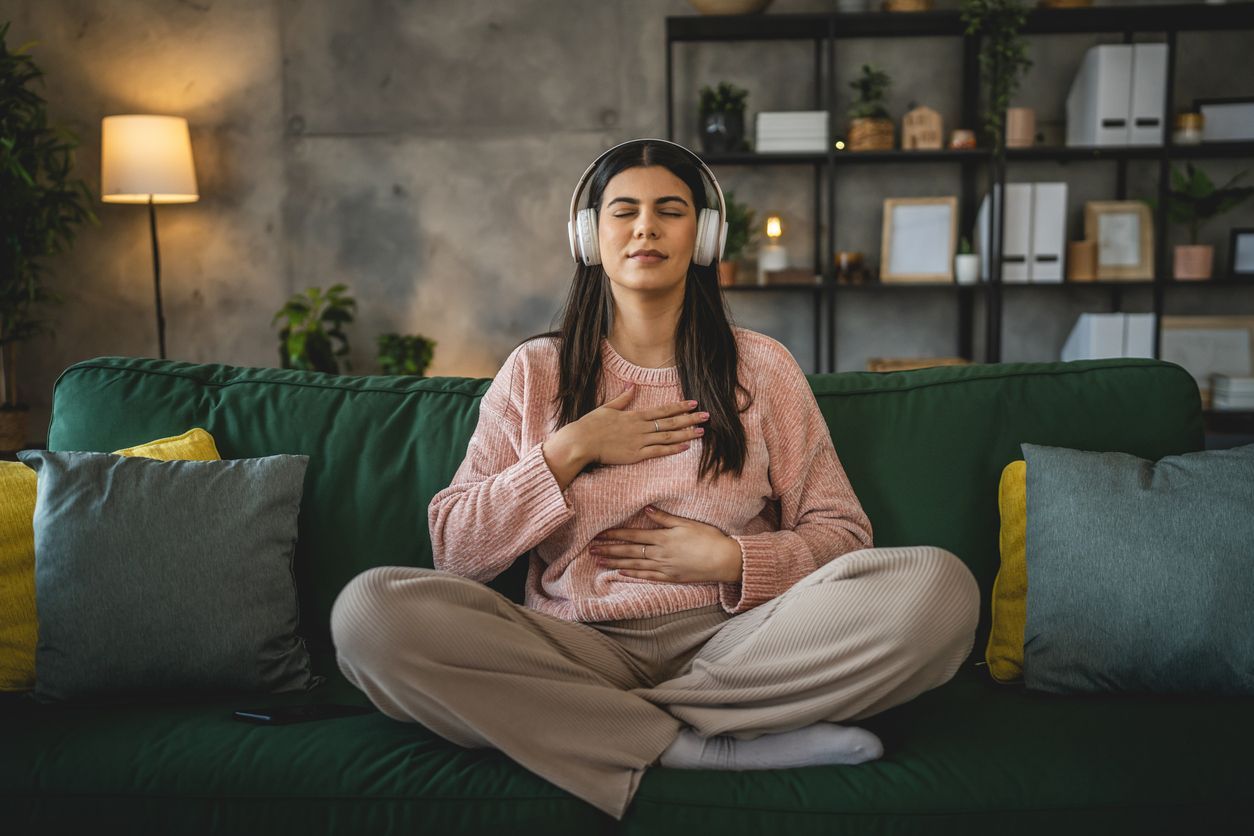 Woman with headphones meditates on a green couch. Hands on chest and stomach; eyes closed.