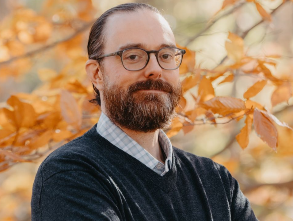 A man with a beard and glasses is standing in front of a tree.