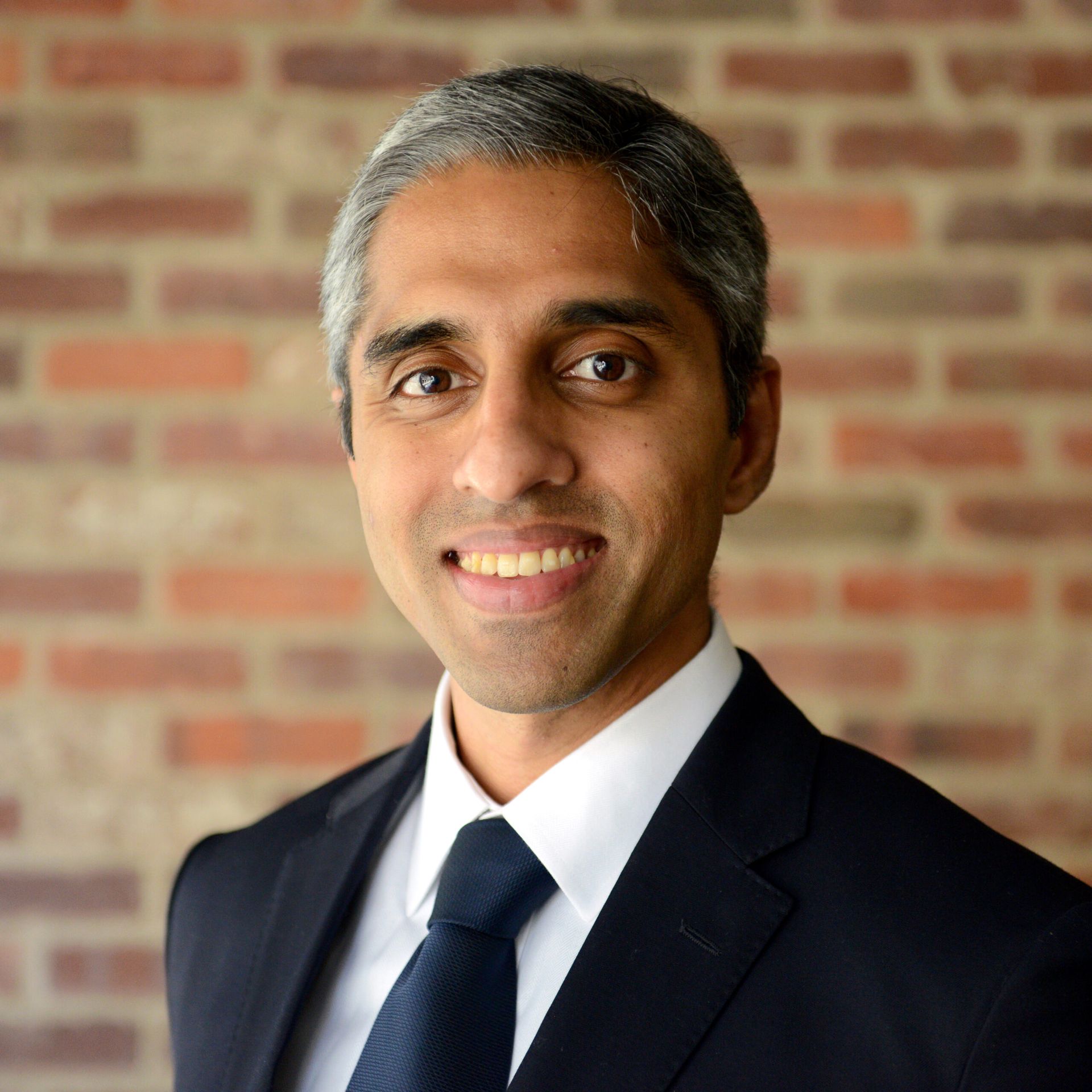 A man in a suit and tie is smiling in front of a brick wall.