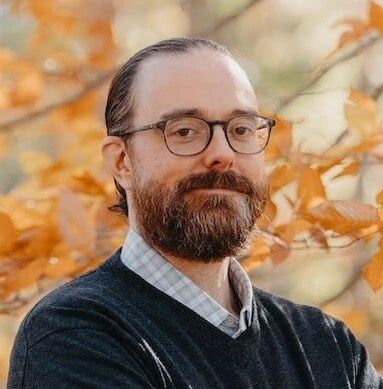 A man with a beard and glasses is standing in front of a tree.