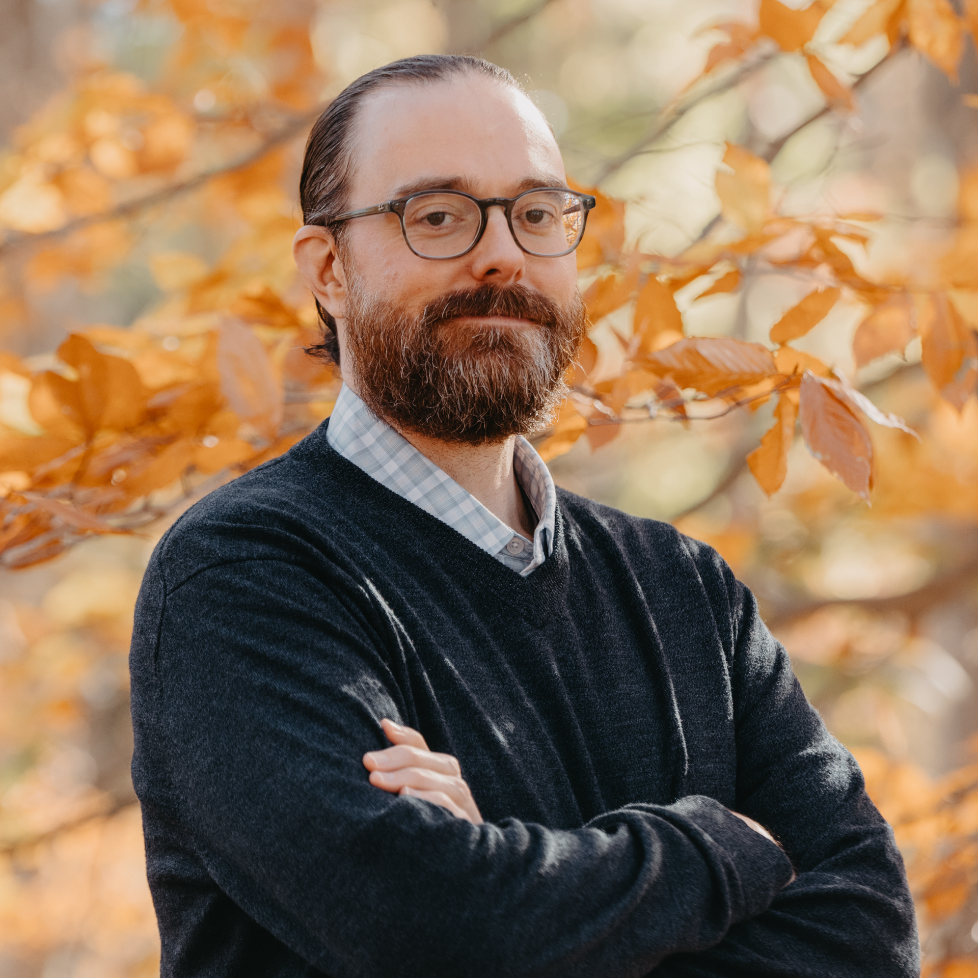 A man with a beard and glasses is standing in front of a tree.