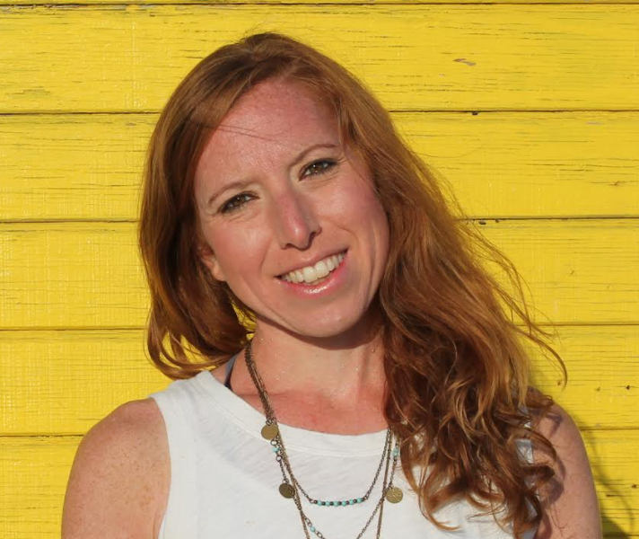 A woman with red hair is smiling in front of a yellow wall