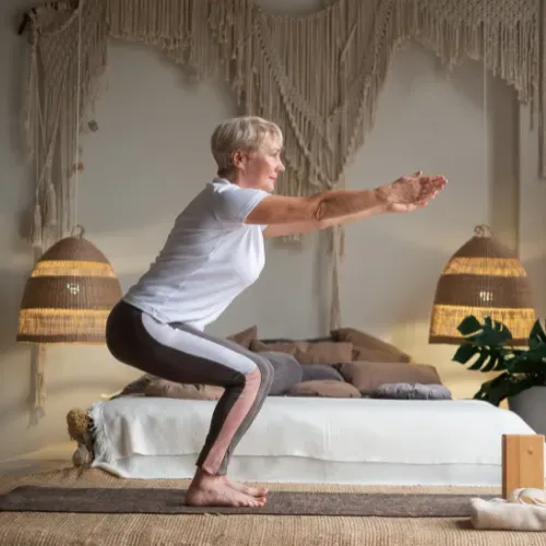 A woman is squatting on a yoga mat in a bedroom.