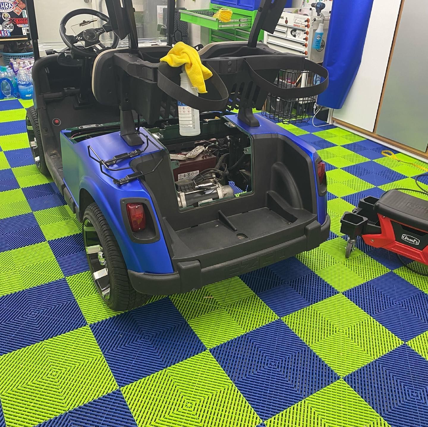Blue golf cart, disassembled, on checkered blue and green floor. Yellow towel. Workshop setting.
