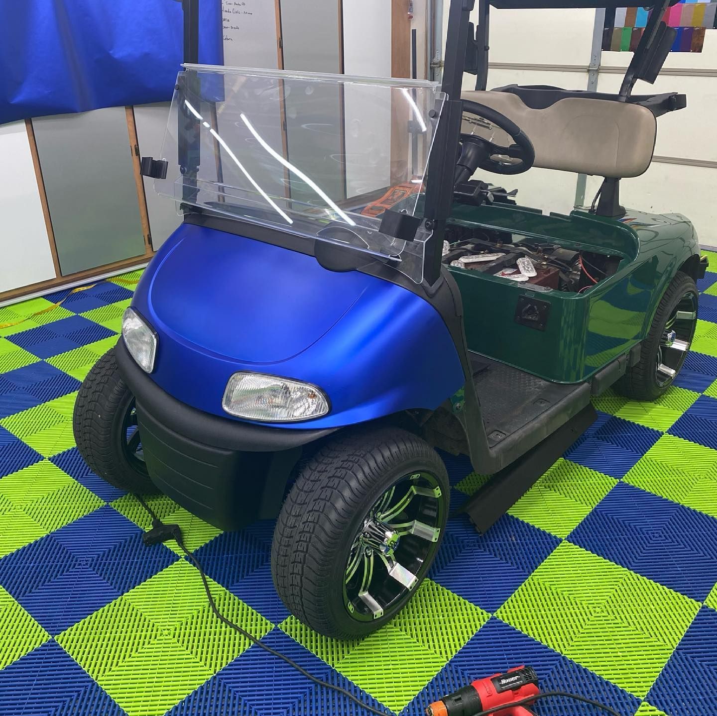 Blue and green golf cart with custom wheels, parked on a checkered floor; heat gun is visible.