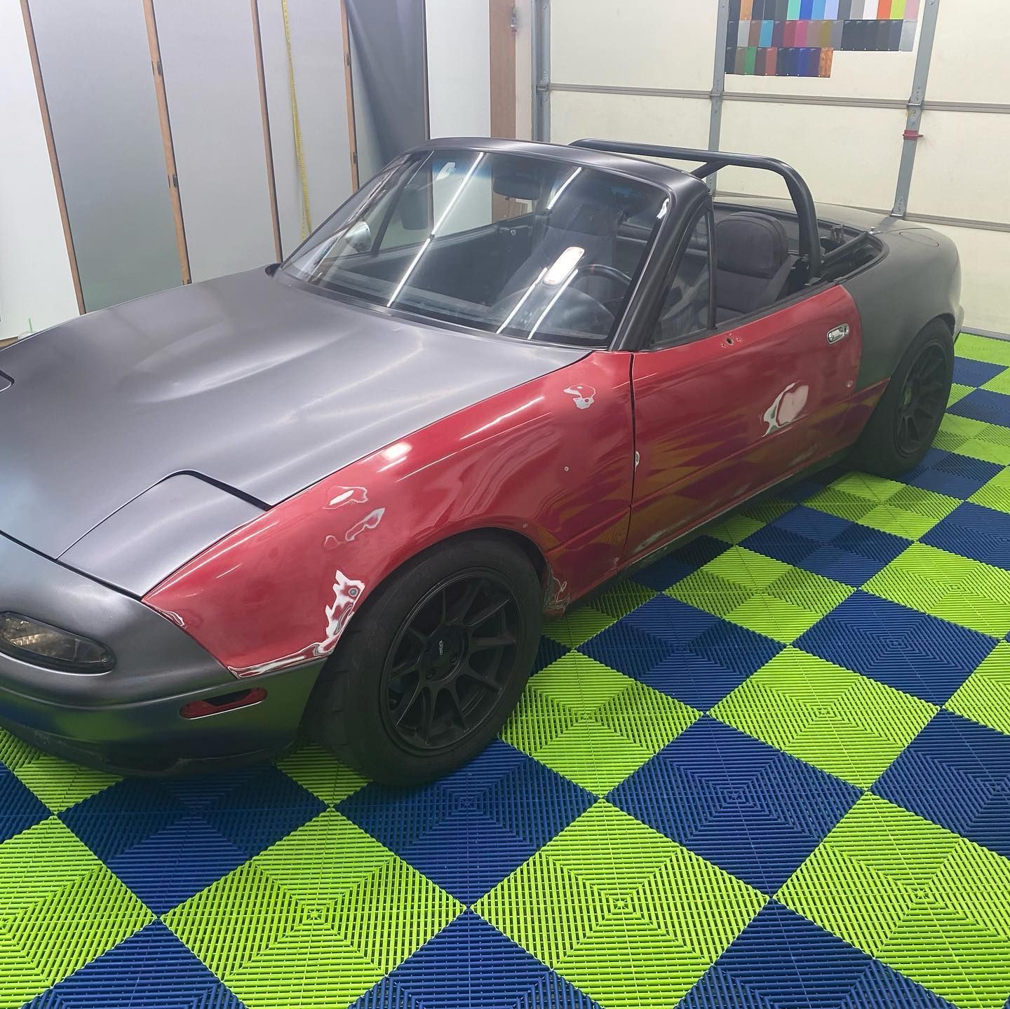 A partially repainted, red and gray convertible car with black wheels, in a garage with checkerboard flooring.