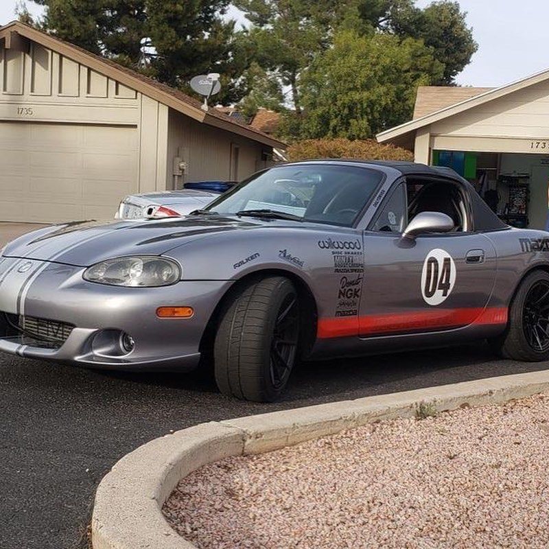 Gray Mazda Miata sports car with racing stripes and number 04 parked in a driveway.