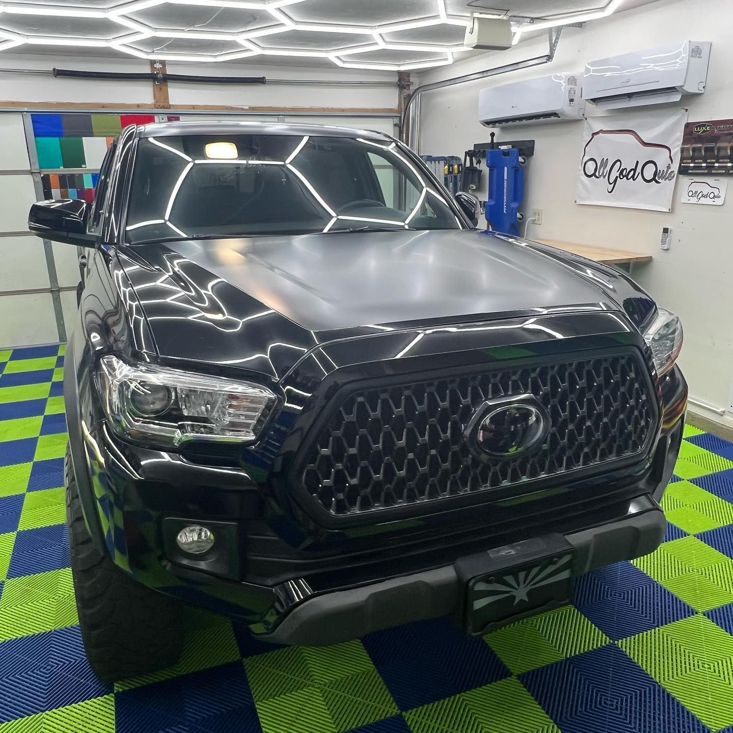 Black Toyota Tacoma truck with a gray hood, inside a well-lit shop with honeycomb lights.