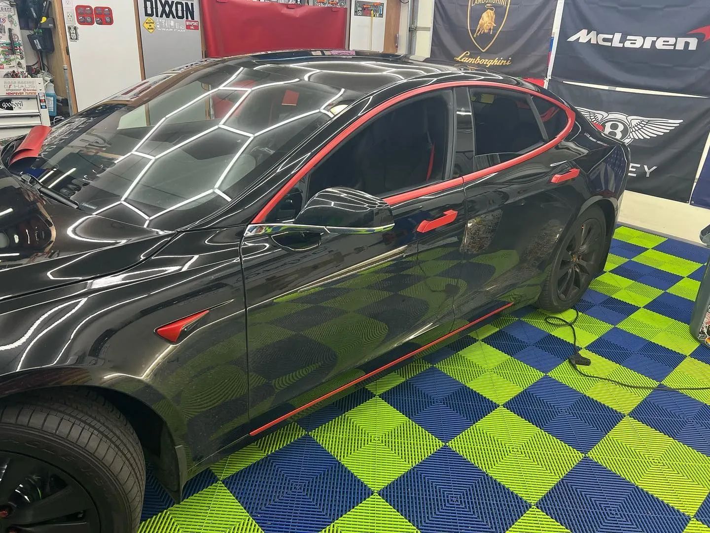 Black Tesla with red accents, parked on a blue and green tiled floor, near flags.