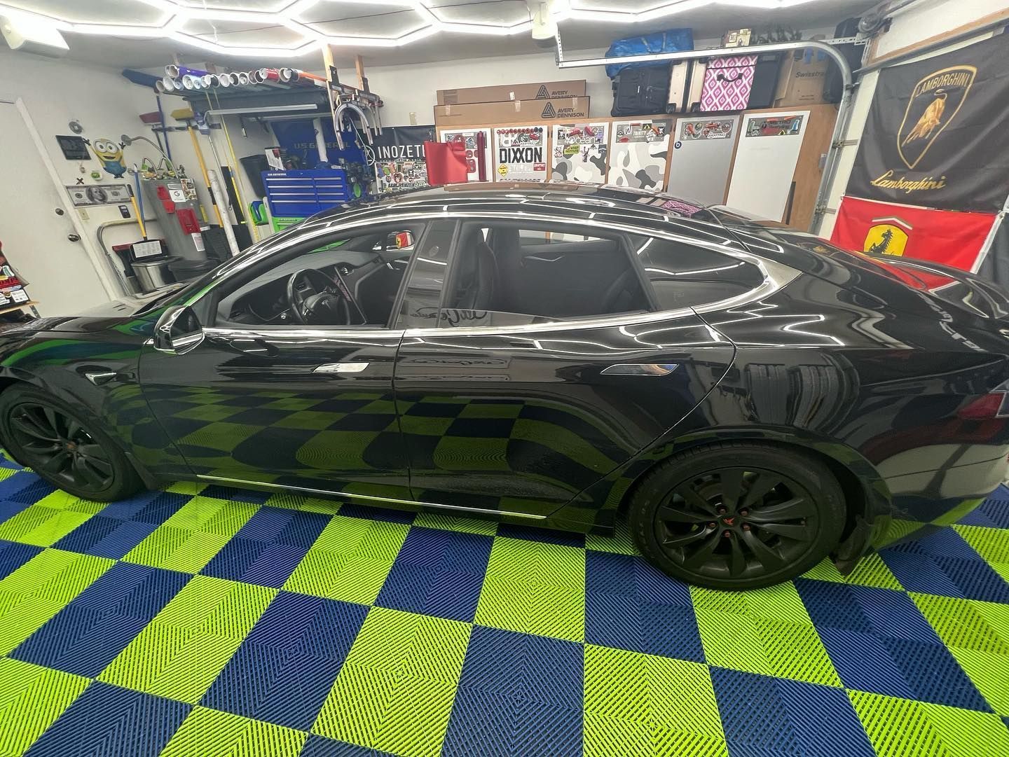 Black Tesla Model S on a checkered blue and green floor, parked in a garage with tools and flags.