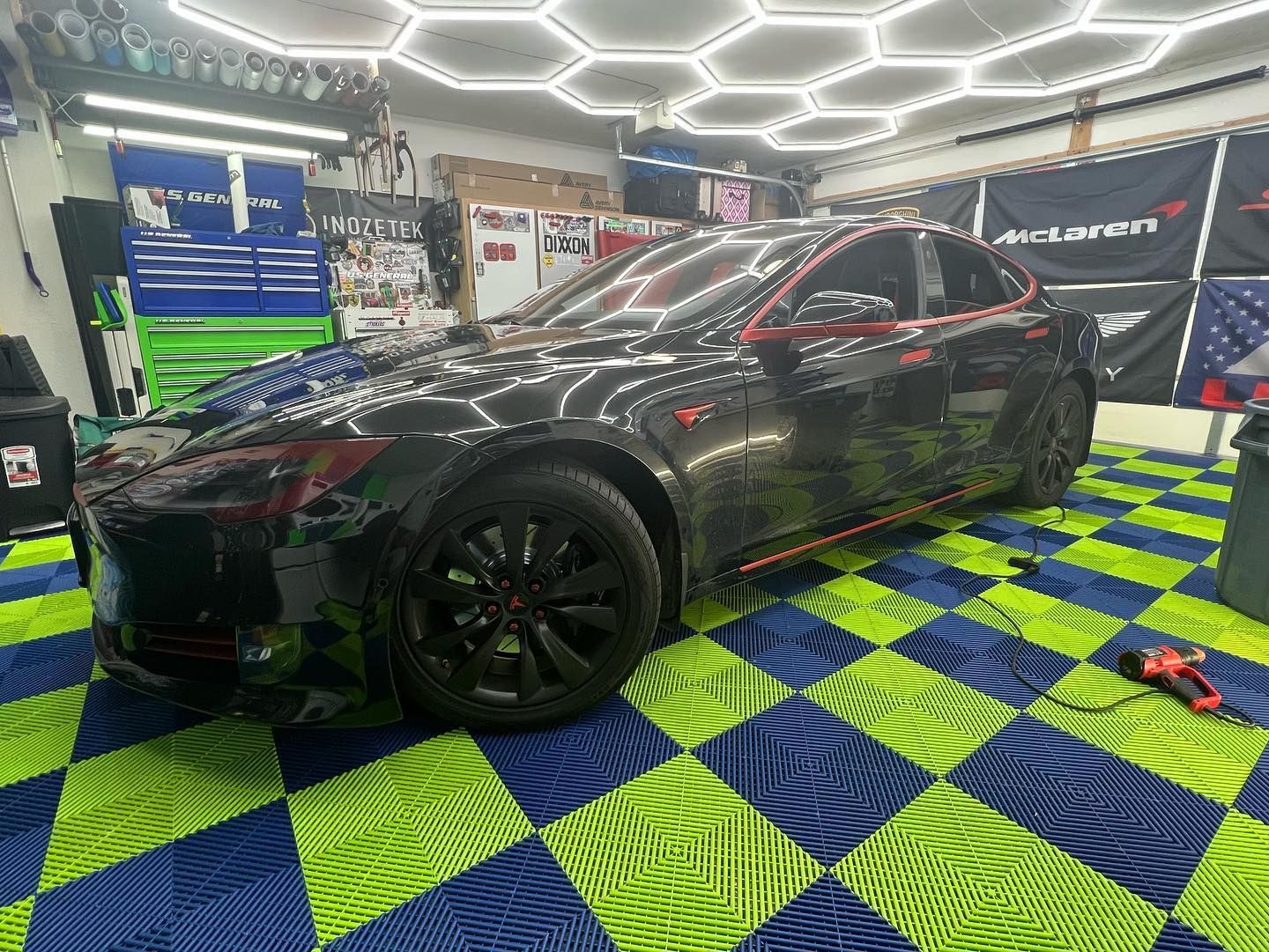 Black Tesla in a garage with red accents, on checkered blue and green floor.