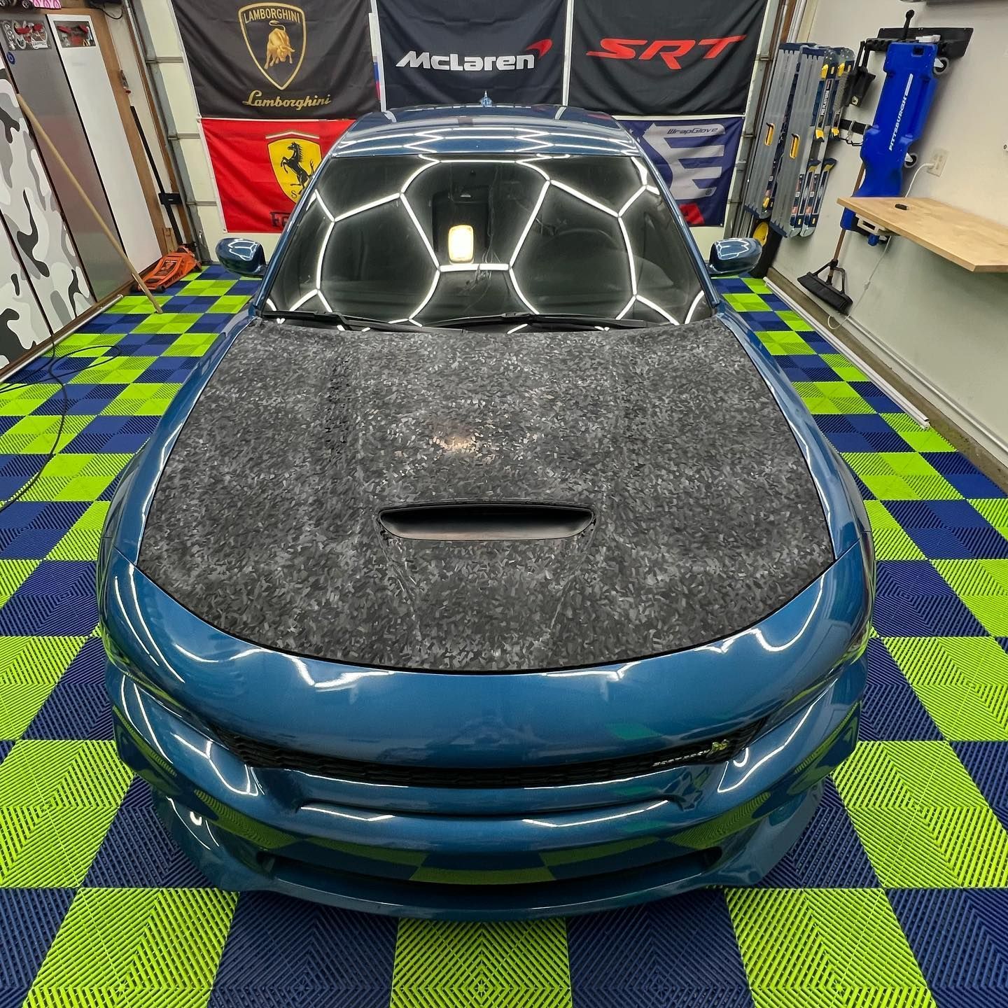 Blue Dodge Charger with a patterned hood sits in a garage with a checkered floor and brand flags.