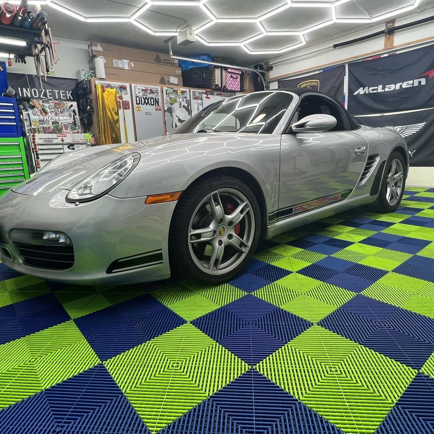 Silver Porsche Boxster convertible on a checkered blue and green floor in a well-lit garage.